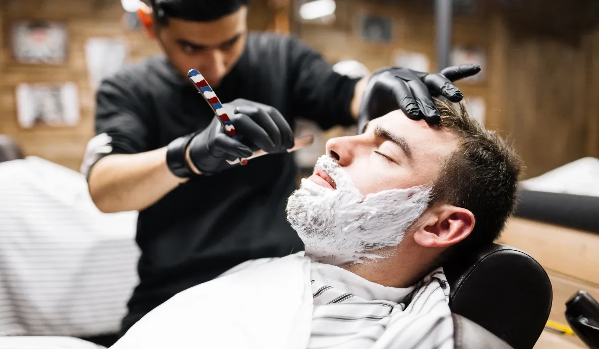 Barbero con guantes negros afeita a navaja a un cliente con espuma