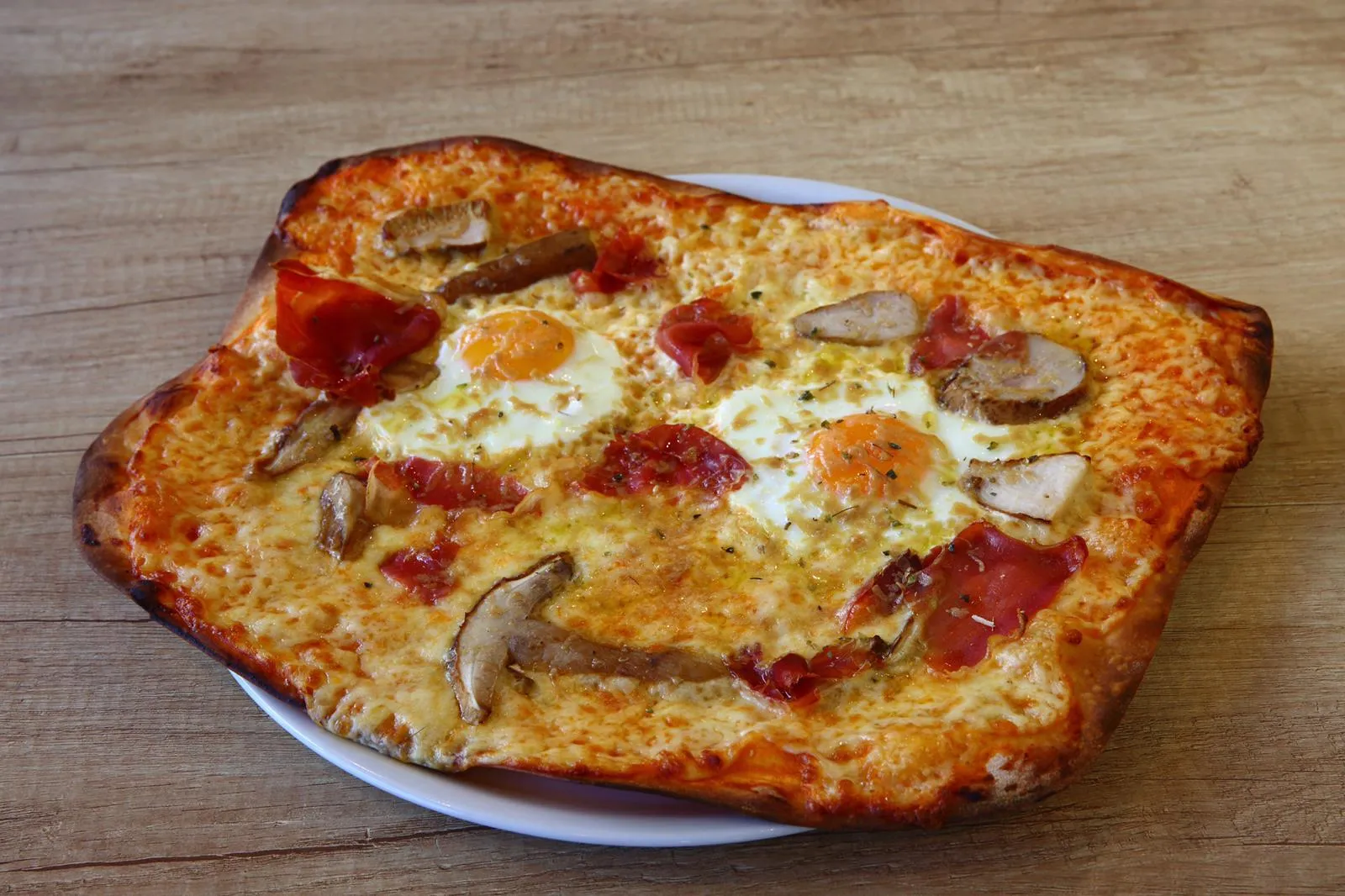 Pizza casera rectangular con huevo, jamón y hongos sobre una mesa de madera