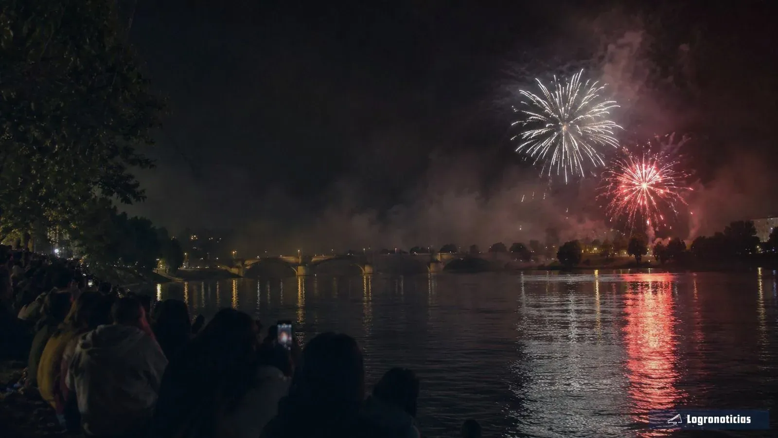Fuegos artificiales sobre el río con su reflejo y público mirando desde la orilla