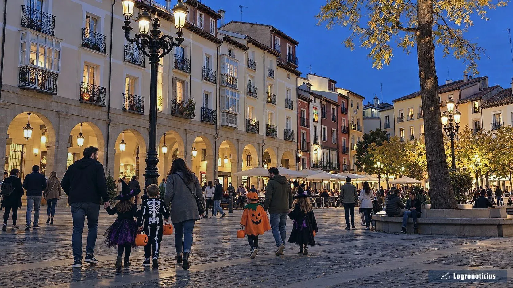 Familias con niños disfrazados cruzando una plaza con soportales al caer la noche