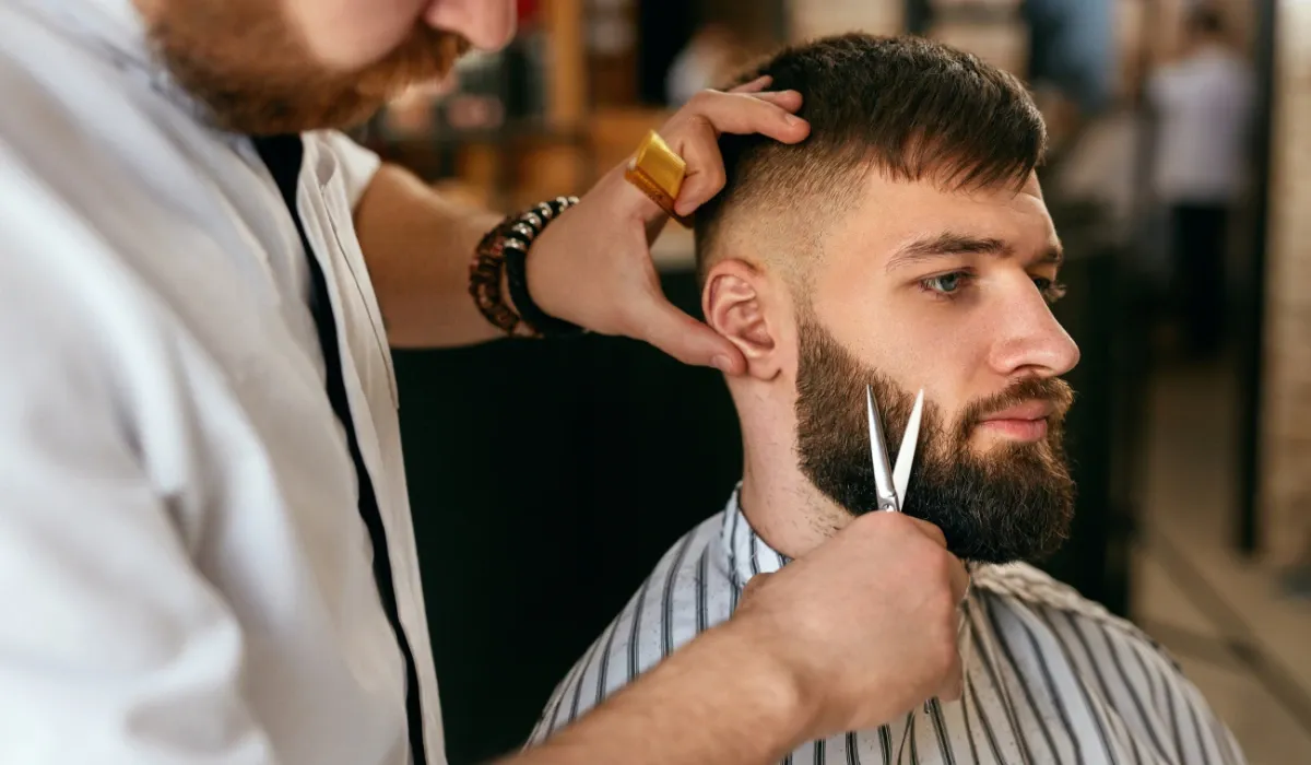 Barbero repasa con tijera la barba de un cliente sentado